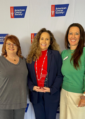 three women standing in front of a American Cancer Society backdrop, middle lady is holding her award